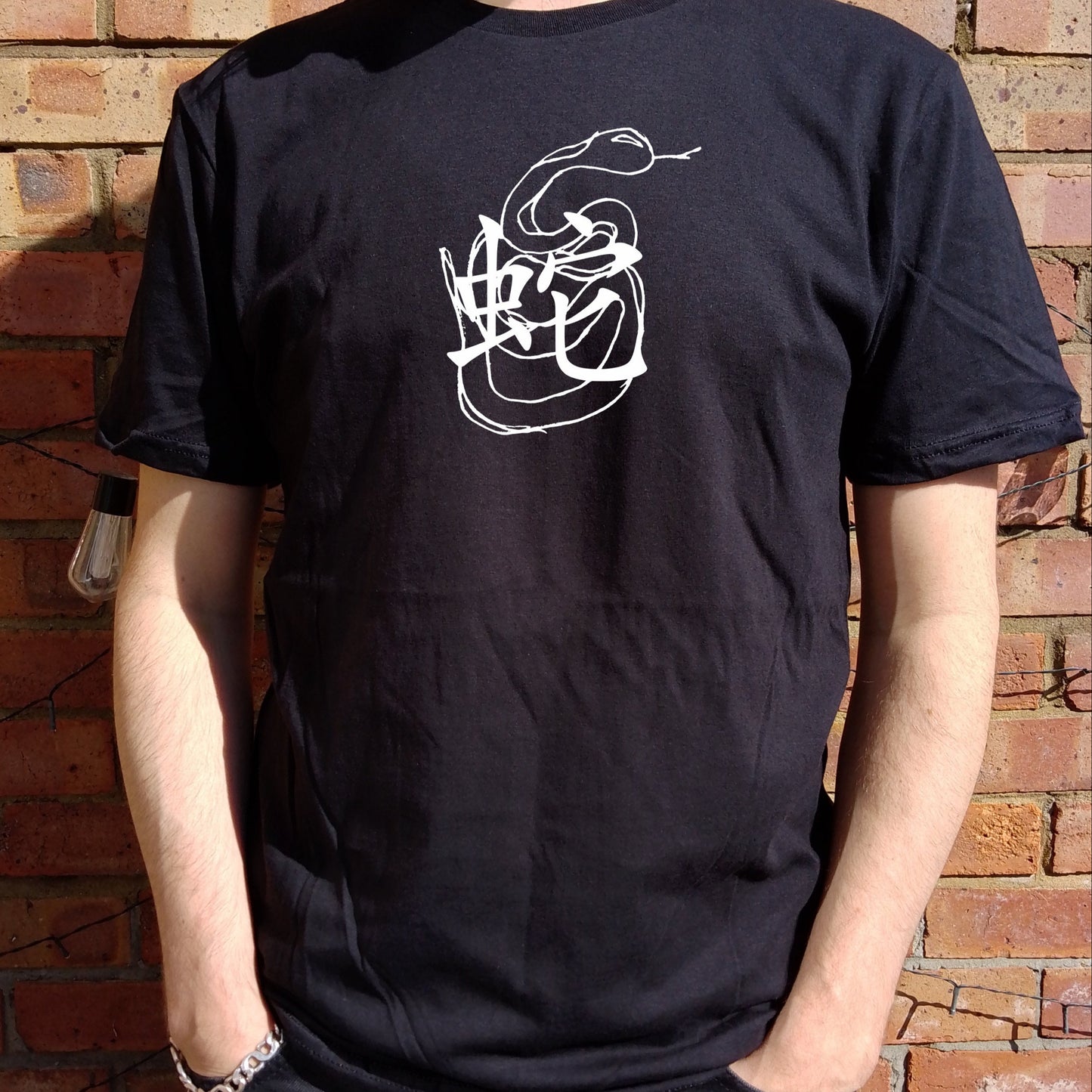 A person wearing a black T-shirt with a white snake symbol design on the front, standing in front of a brick wall.