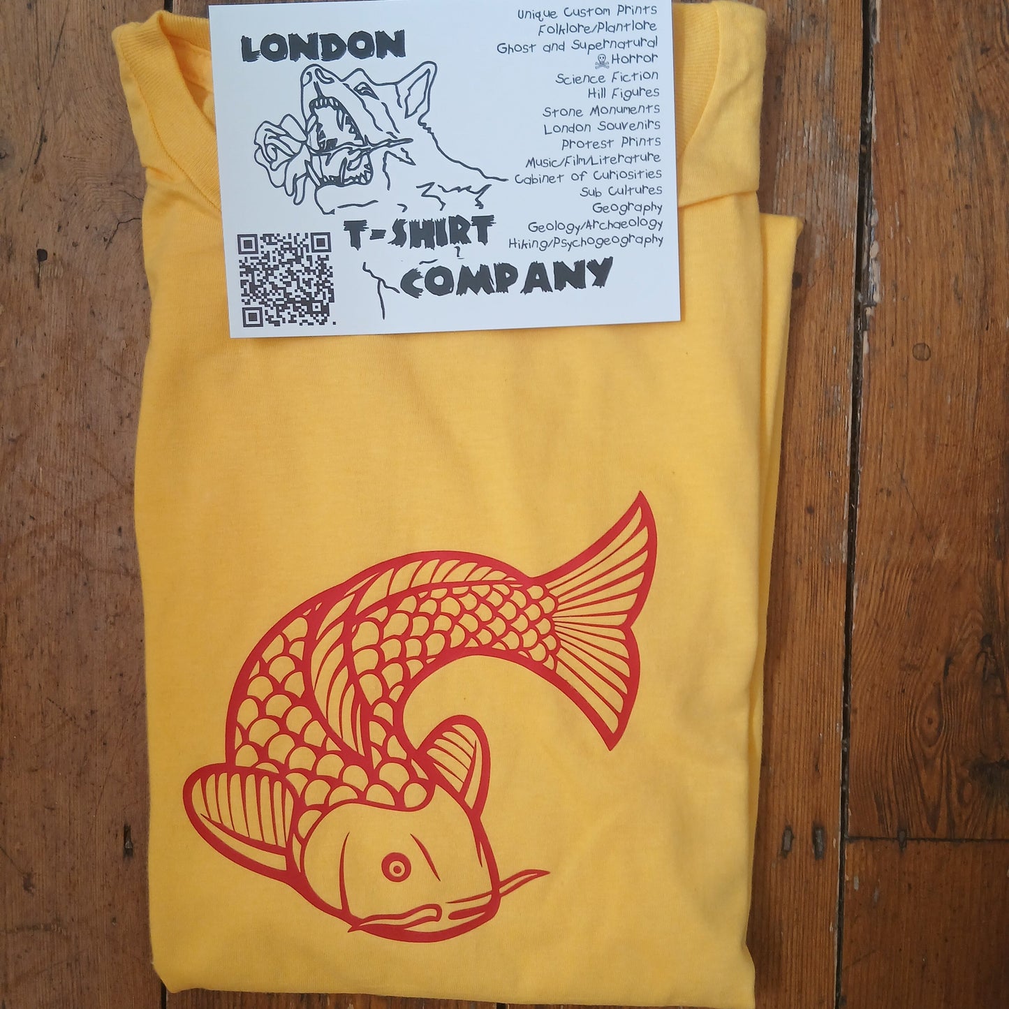 A folded yellow T-shirt laid flat on a rustic wooden surface, featuring a large red koi fish graphic on the front. A white paper label card with the brand name 'T-Shirt Company' and a QR code is placed on top of the shirt.